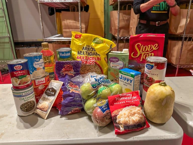 A variety of nonperishable and fresh food items are arranged on a table, including canned goods like turkey gravy, green beans, evaporated milk, and cranberry sauce, as well as pantry staples like stuffing mix, corn muffin mix, instant mashed potatoes, and vegetable oil. Also shown are fresh apples, a butternut squash, a pack of multigrain cereal, cocoa mix, and fruit snacks. In the background, paper grocery bags on metal shelving for Food For All DC recipients.