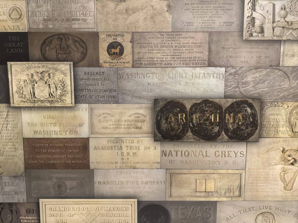 A collage of commemorative stone and metal plaques mounted on a wall, honoring various states, organizations, and individuals for their contributions to the Washington Monument. Prominent plaques include engraved names like "ARIZONA" and dedications from places such as Virginia, Texas, and the City of New York.