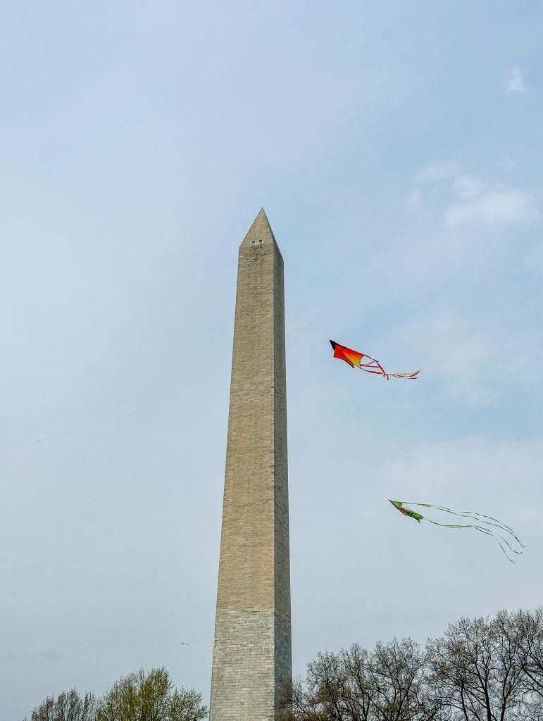 Playful springtime image of the Washington Monument with colorful kites flying in the sky above. The monument towers over the scene, with trees in the foreground and a light, overcast sky, capturing the festive and airy mood of the moment.