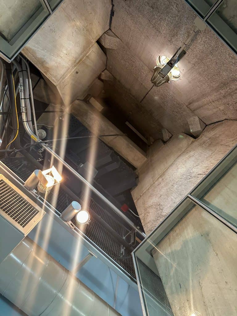 An upward view inside the Washington Monument’s elevator shaft, showcasing rugged stone walls, steel framework, and bright spotlights illuminating the structure's interior.