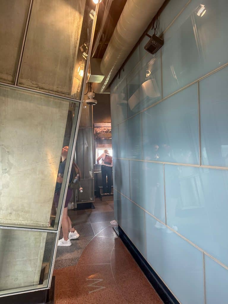 Interior view of the Washington Monument observation deck with slanted glass panels, smooth flooring with a directional line and “W” marking, and visitors looking out from viewing windows at the far end.