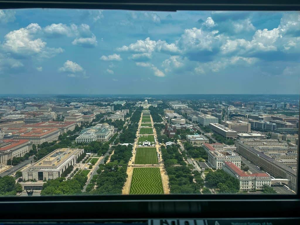 Aerial view from the top of the Washington Monument looking west down the National Mall, with the U.S. Capitol in the distance, flanked by rows of federal buildings and museums, under a bright sky with scattered clouds.