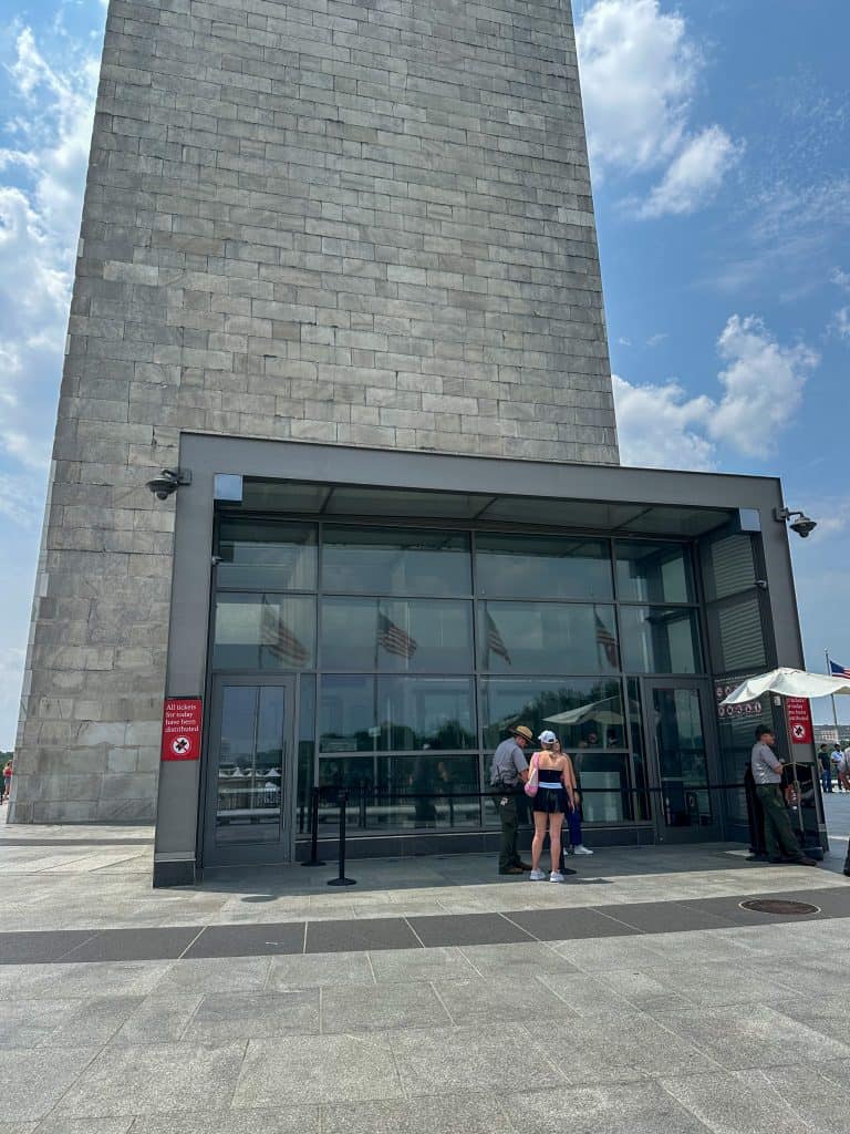 Ground-level view of the base of the Washington Monument showing the modern glass entry pavilion where visitors check in, with two uniformed park rangers standing near the door and a sign stating all tickets have been distributed.