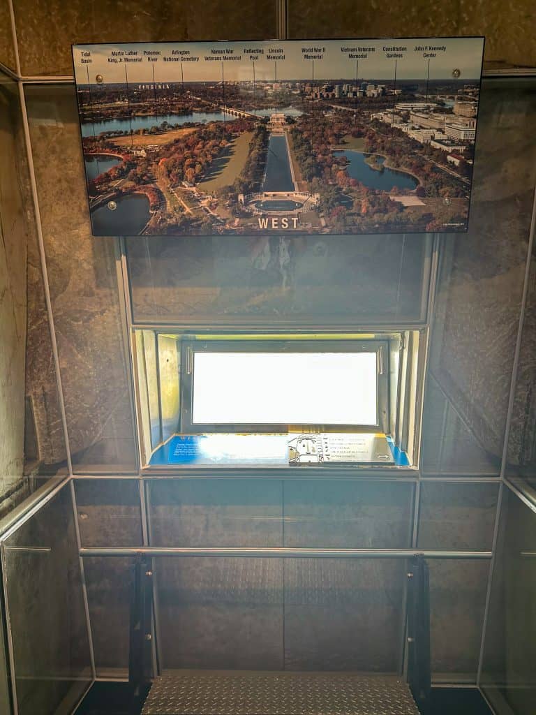 Interior view of a Washington Monument observation window labeled “WEST,” featuring a backlit directional display above the window that identifies landmarks such as the Lincoln Memorial, Reflecting Pool, and Arlington National Cemetery. A small viewing platform with a metal floor and handrails allows visitors to look out over the western landscape of Washington, D.C.