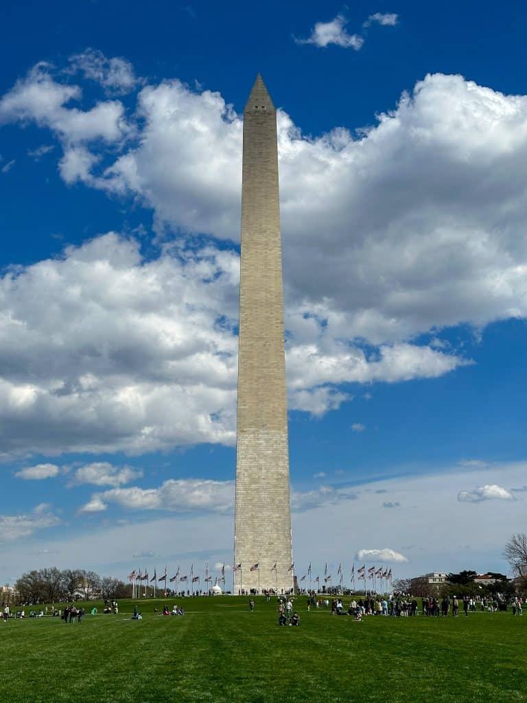 Daytime shots of the Washington Monument under a bright blue sky, with fluffy white clouds in the background. The monument stands tall, surrounded by a green lawn where people are gathered, creating a picturesque view of this national landmark.
