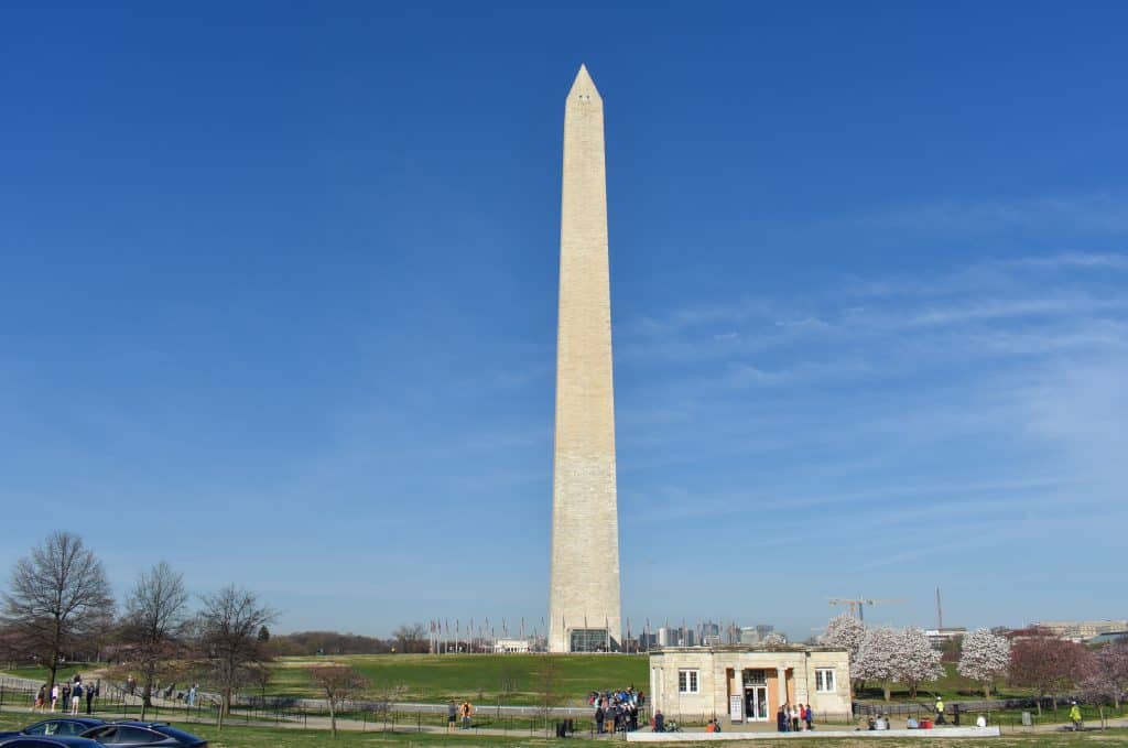 A bright daytime view of the Washington Monument standing tall and surrounded by a lush green lawn. In the foreground, small groups of people are walking or resting, while the monument’s entrance building and the circle of American flags at its base are visible. The sky above is clear and bright blue, adding to the grandeur and openness of the scene.