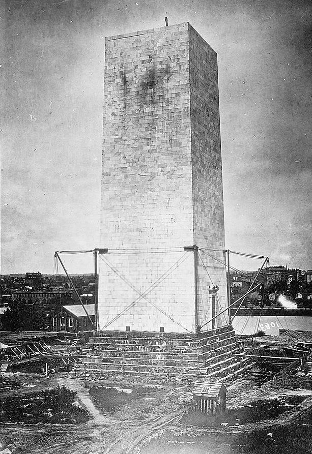 Black-and-white photograph of the Washington Monument during construction, with the uncompleted stone obelisk standing starkly against the sky. Metal scaffolding supports the partially built structure, and stone blocks and building materials are scattered on the ground below. A person stands atop the monument, silhouetted against the sky. The surrounding landscape includes a few small buildings and dirt paths, hinting at the period's less-developed setting.
