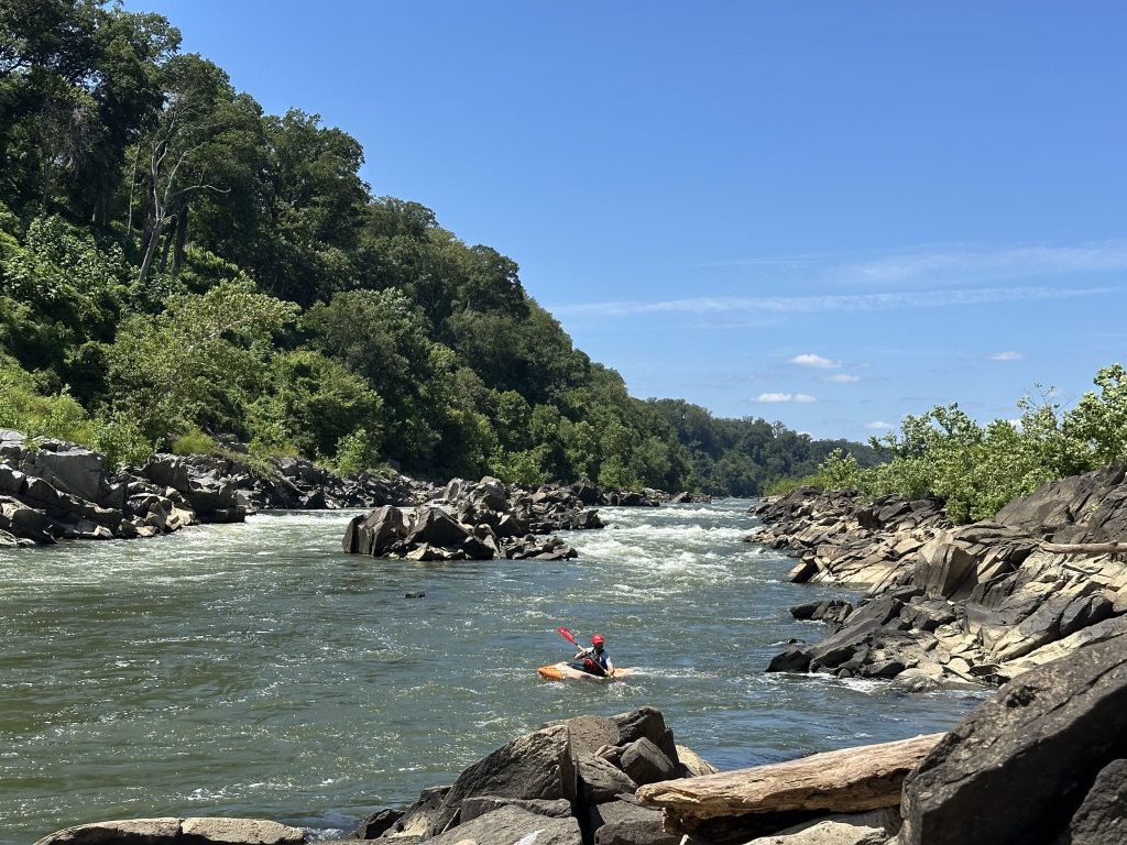 Solo kayaker paddling through a rocky section of the Potomac River with Little Falls in the background under a clear blue sky, surrounded by lush green trees and rugged shoreline.