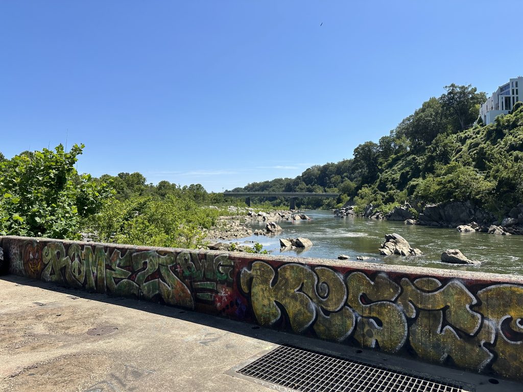 Graffiti-covered concrete wall overlooking a rocky section of the Potomac River, with green hills, scattered boulders, and a bridge visible in the distance under a clear blue sky.
