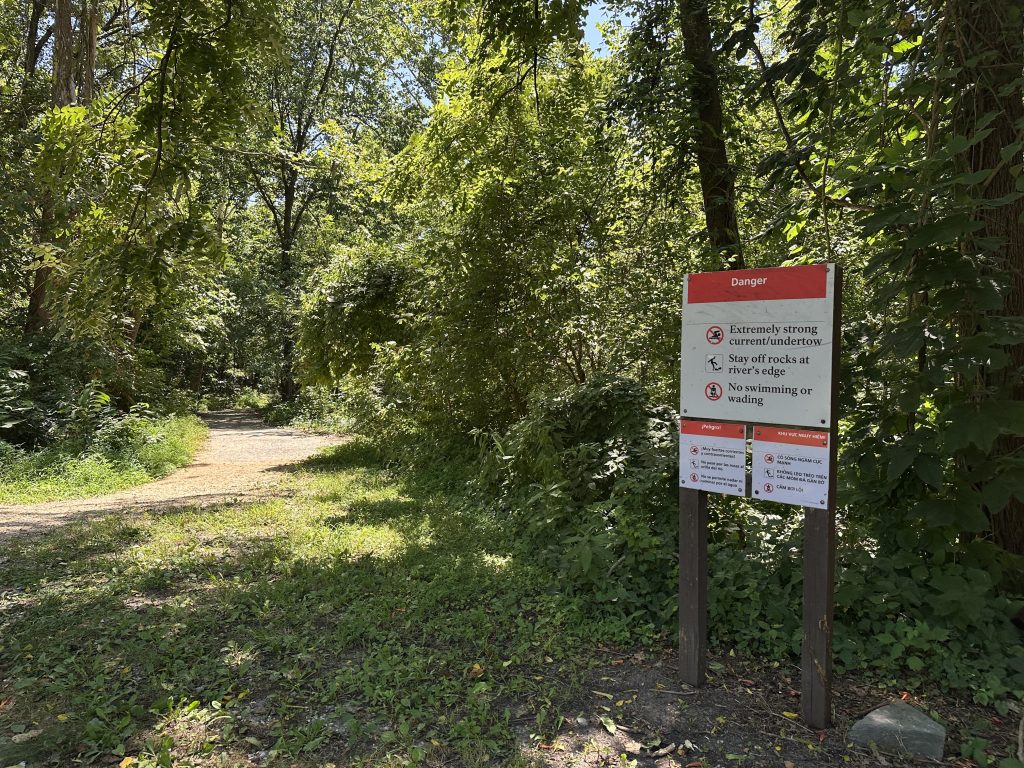 Warning sign at the entrance of a wooded trail, cautioning visitors about extremely strong currents, dangerous rocks, and prohibiting swimming or wading near the river.