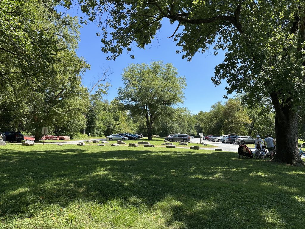 Sunny park scene with scattered cars in a tree-lined parking lot, grassy picnic area in the foreground, and two cyclists with a child trailer resting under the shade.
