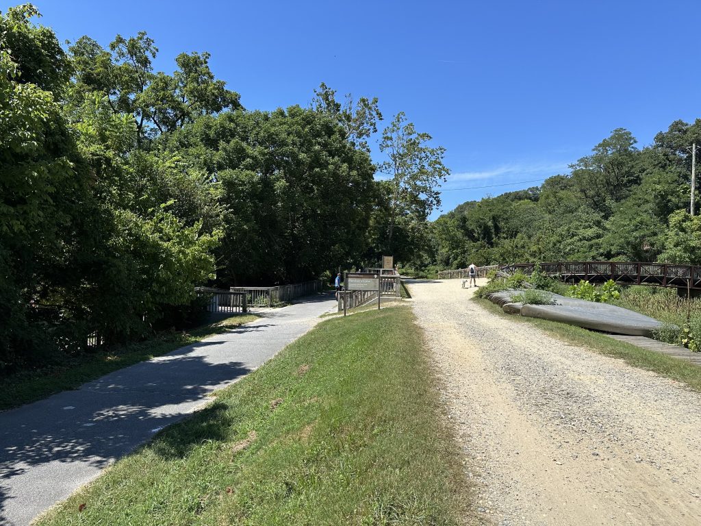 Split trail with a paved path and gravel path running through a wooded area under a clear blue sky, with canoes stacked beside the trail and a cyclist in the distance.