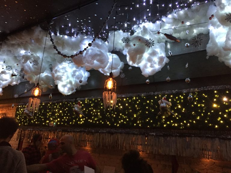 Dimly lit bar interior decorated with glowing cotton clouds, snowflakes, and hanging ornaments to create a winter wonderland effect. Icicle-style light fixtures hang from the ceiling, while fairy lights and festive figures like Santa-riding-narwhals line the wall above the bar.